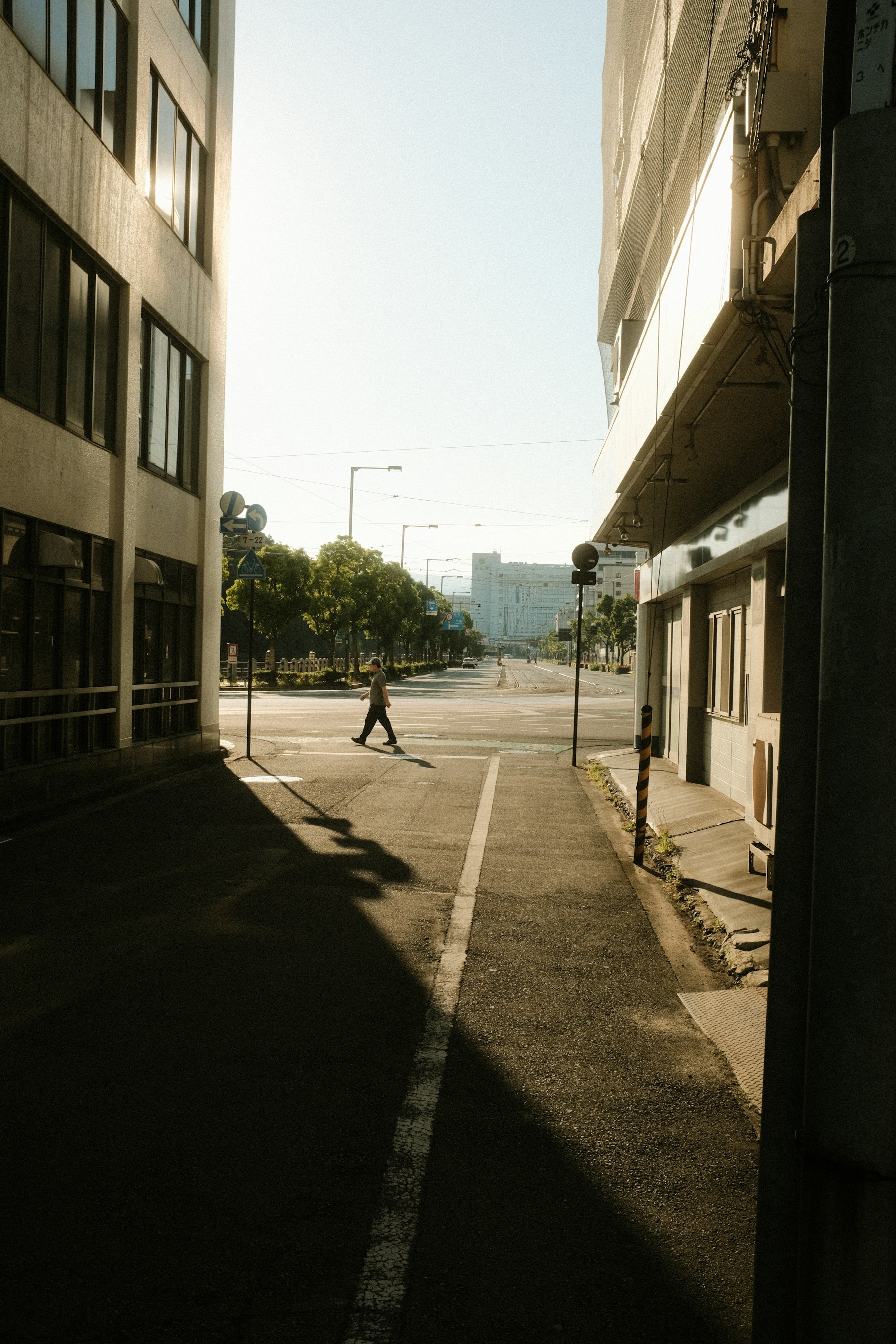 A lone pedestrian crossing a sunlit street between deep building shadows