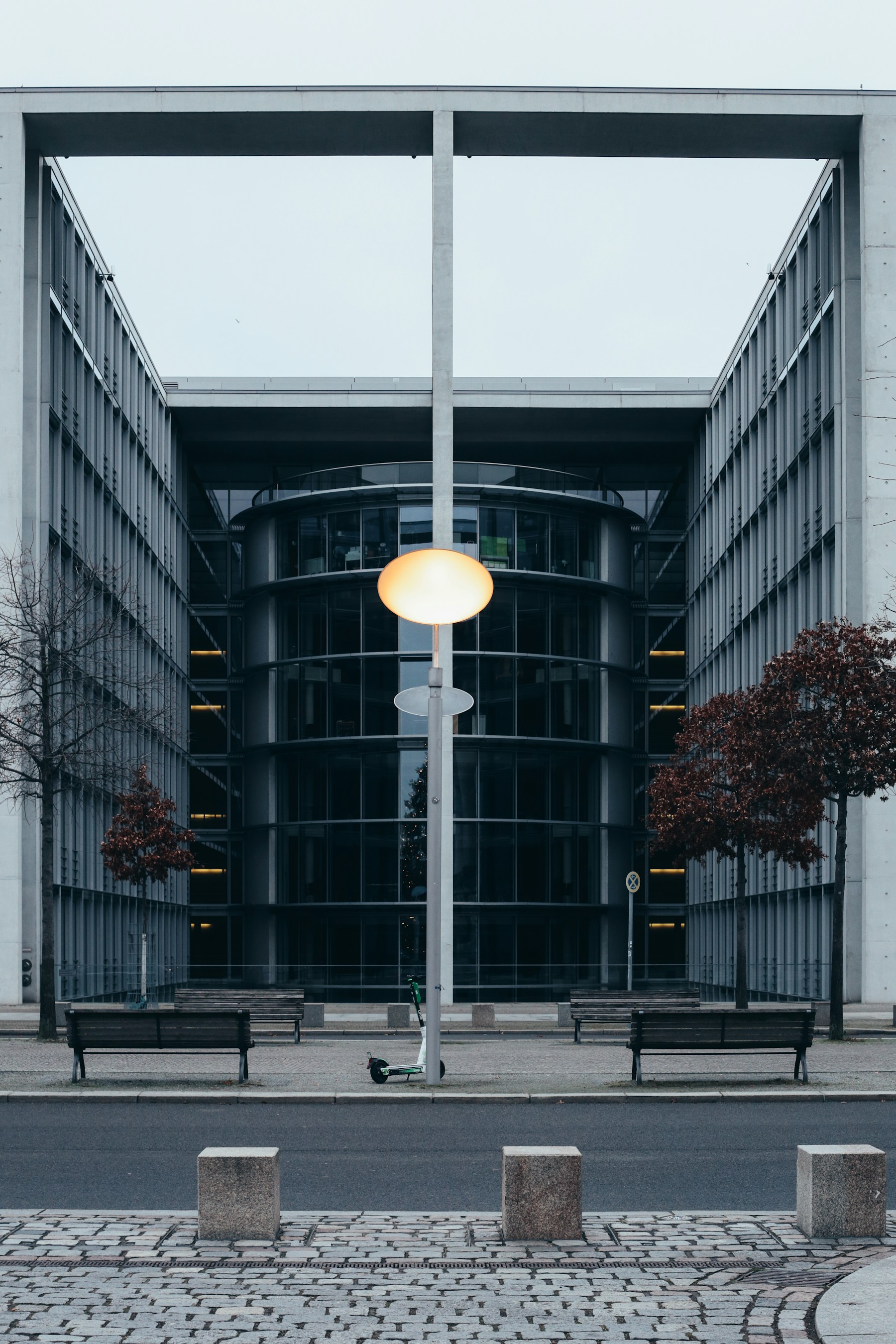A symmetrical concrete facade with benches, bollards, and a glowing street lamp